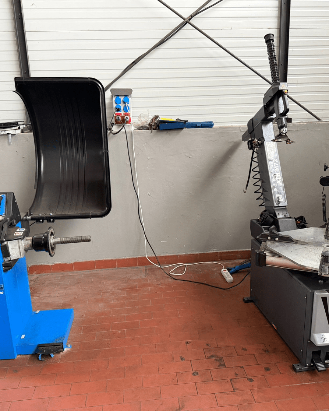 Auto shop equipment: a blue tire changer on the left and a vertical wheel balancer on the right in a workshop with red tile floor and cords strewn about.