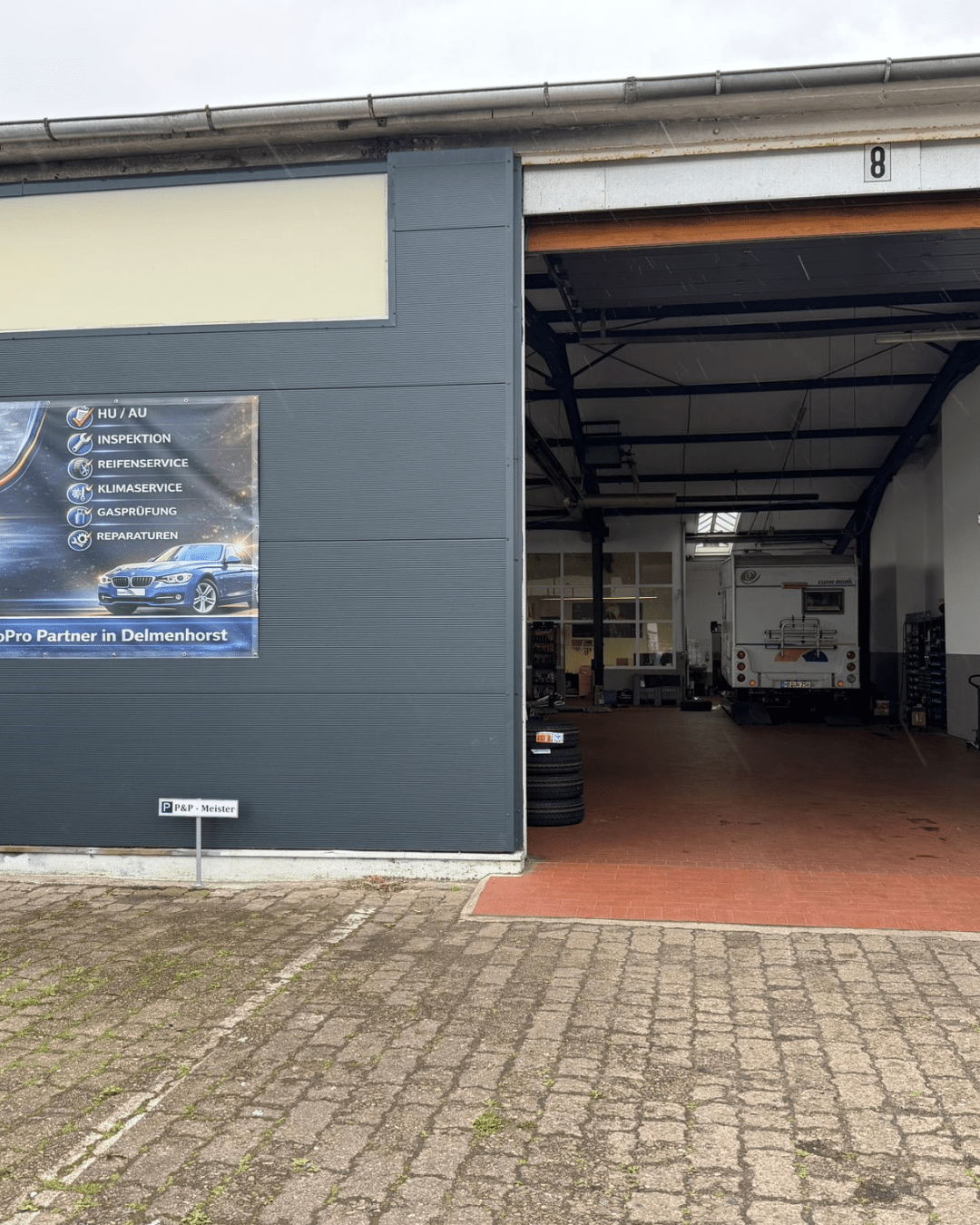 Open automotive repair bay with a large white vehicle inside, dark gray exterior walls, and a service sign on the wall to the left.